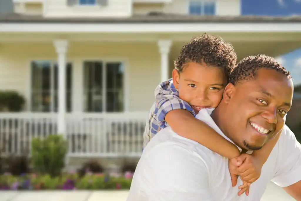 A smiling man carries a young boy on his back in front of a house with a porch and white railing. Both look happy and are outdoors on a sunny day.
