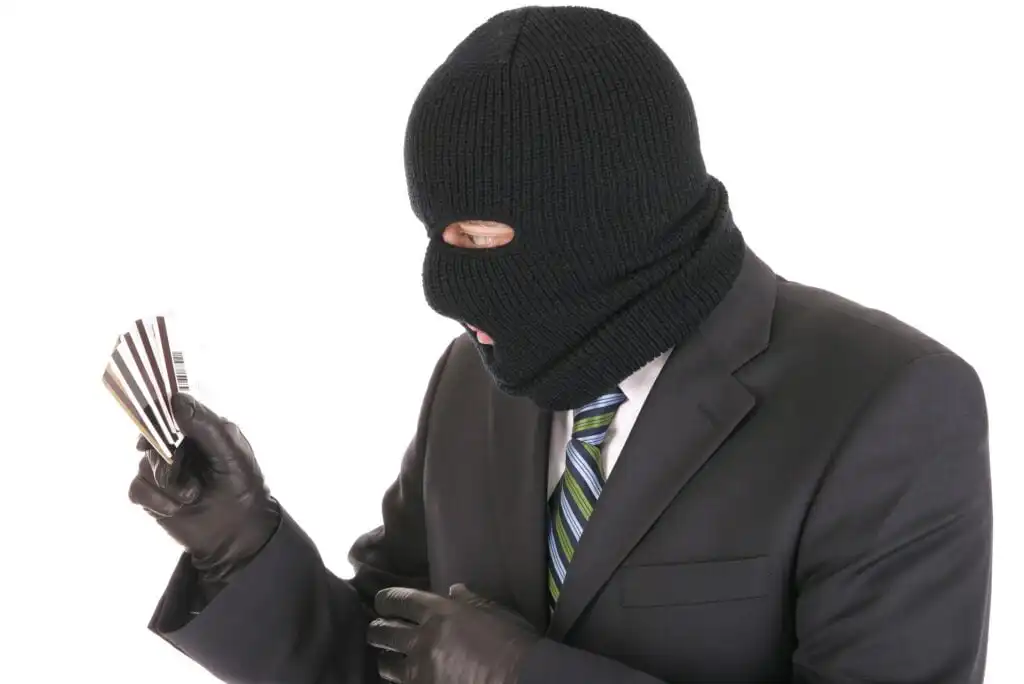 A person in a black ski mask, suit, and gloves holds several credit cards while looking at them; the background is white.