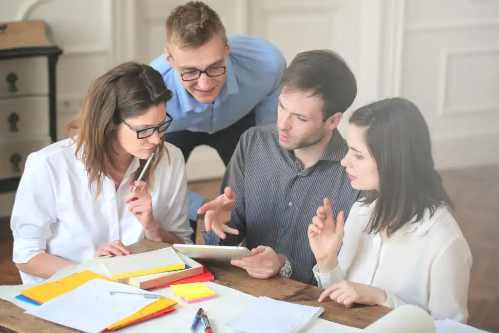 Four people sit and stand around a table with notebooks and papers, closely collaborating and discussing something on a tablet. They appear focused and engaged in a work or study environment.