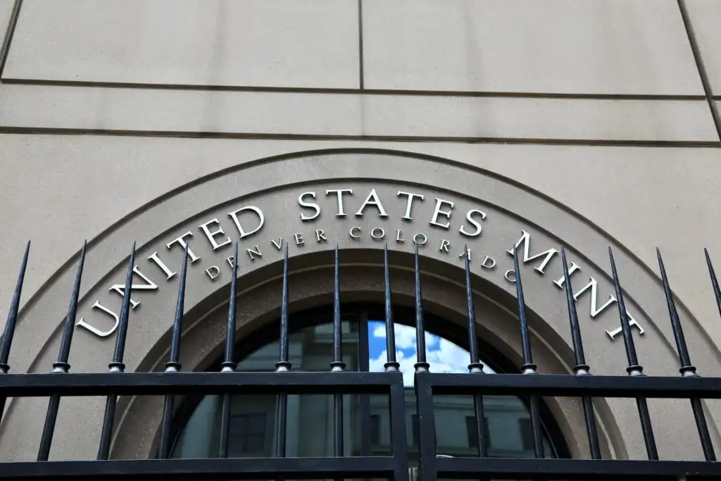 The entrance to the United States Mint in Denver, Colorado, showing an arched window with the words United States Mint above and an iron fence in the foreground.