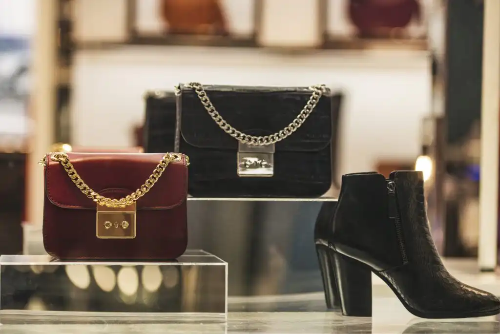 Three luxury handbags, two with gold chain straps—one burgundy and one black—and a pair of black heeled ankle boots are displayed on clear stands in a well-lit store.