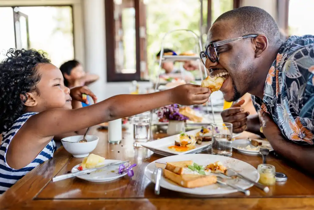 A child in a striped shirt feeds a smiling adult wearing glasses across a table set with plates of food, drinks, and breakfast dishes in a bright, cheerful room.