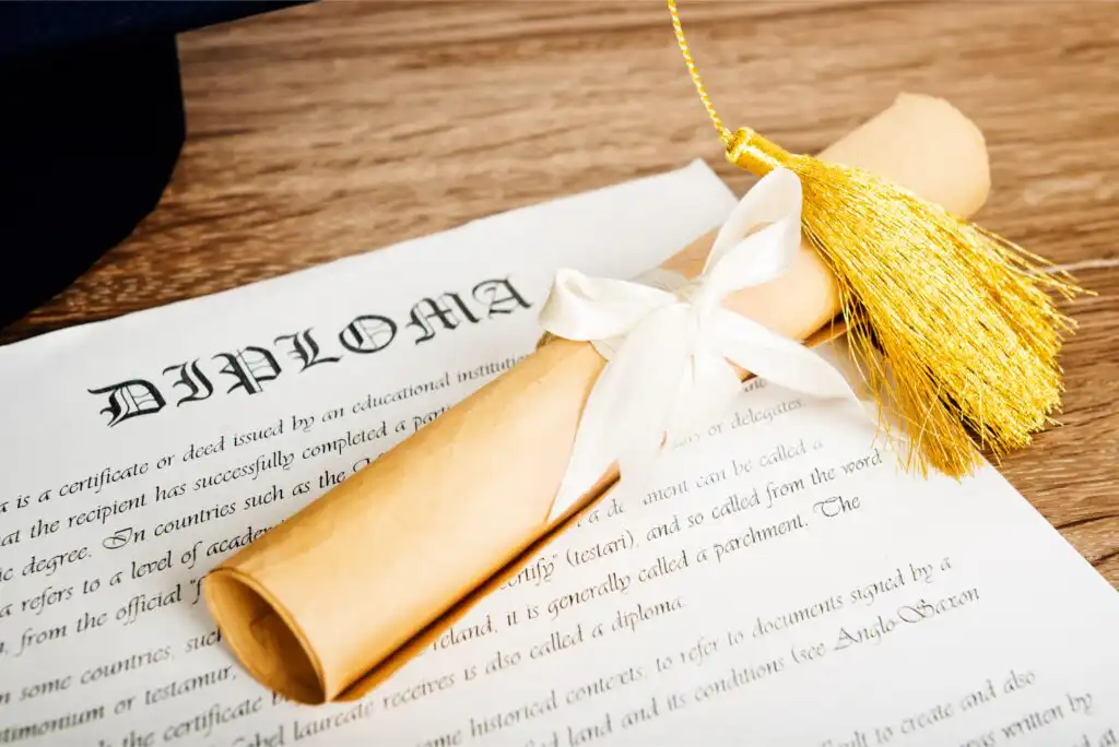 A rolled-up diploma tied with a white ribbon and a gold tassel rests on top of an unrolled diploma certificate on a wooden surface. A graduation cap is partially visible in the background.