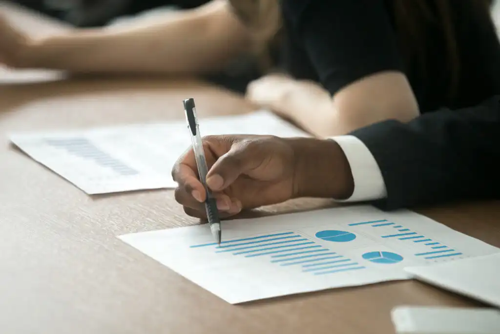 Close-up of a person in business attire holding a pen and reviewing a printed page with blue bar charts and pie graphs at a desk, with another person blurred in the background.