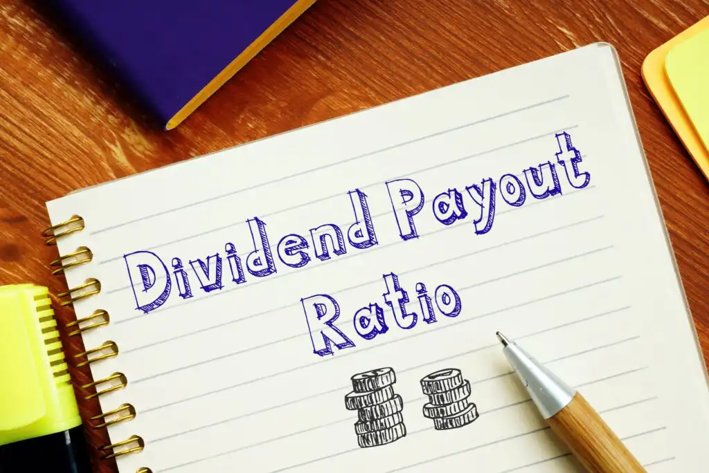A notebook page with “Dividend Payout Ratio” written in blue marker, an illustration of stacked coins, a silver pen, and office supplies like a yellow highlighter and a purple notebook in the background.
