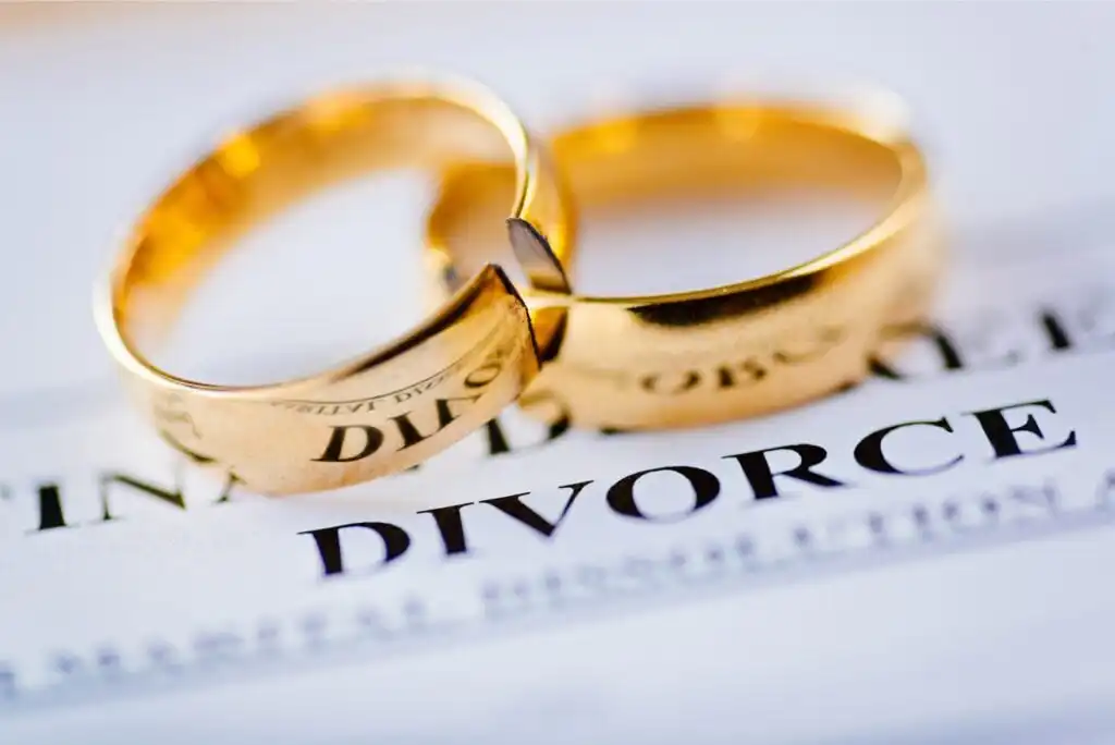 Two gold wedding rings, one of which is broken, rest on a document with the word DIVORCE printed in bold letters, symbolizing the end of a marriage.