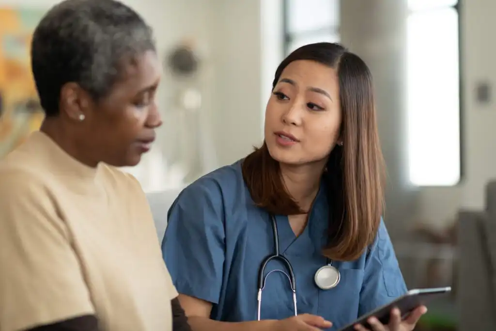 A nurse wearing scrubs and a stethoscope sits beside an older woman, holding a tablet and speaking with her in a caring manner in a bright indoor setting.