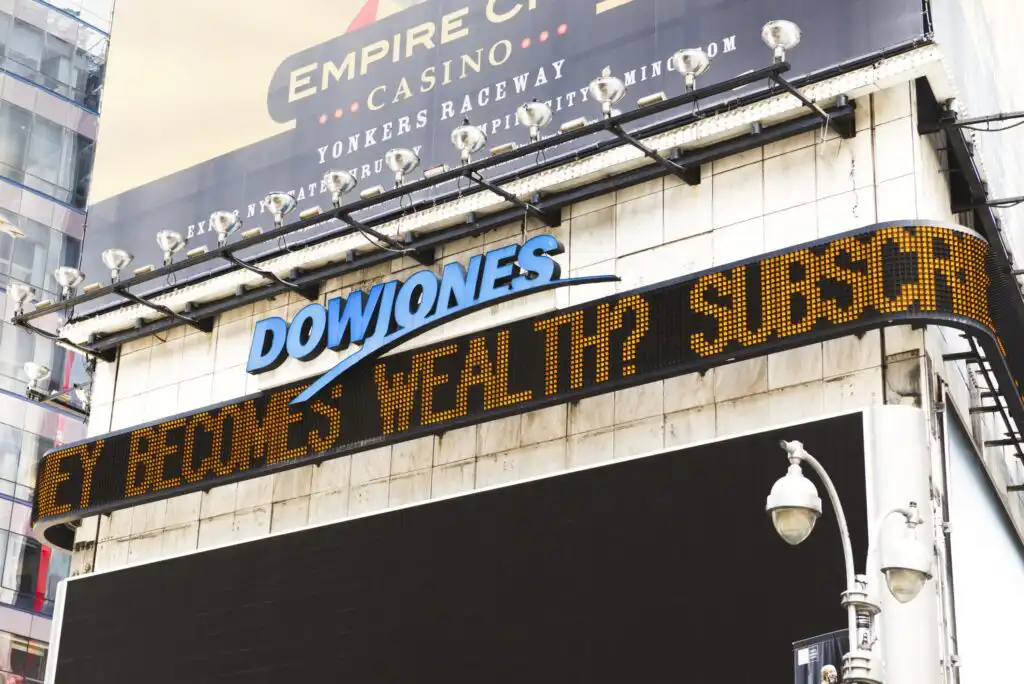 A digital news ticker in Times Square displays “DOW JONES” and scrolling text. A partially visible billboard and a streetlamp are above and below the ticker. The scene is set in an urban environment.