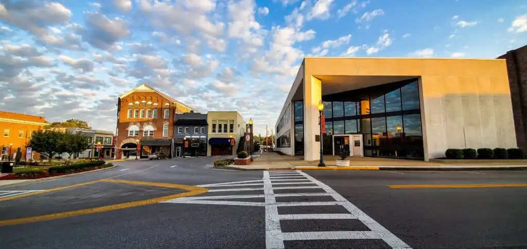 A wide street in a small town with brick and modern buildings, large windows, and lamp posts, under a sky filled with fluffy clouds at sunrise or sunset. Crosswalk lines are visible in the foreground.