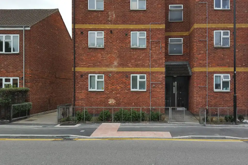 A red brick apartment building with yellow brick lines, white-framed windows, and a dark entrance. A paved sidewalk, small fence, and some green bushes are in front, with a street and curb in the foreground.