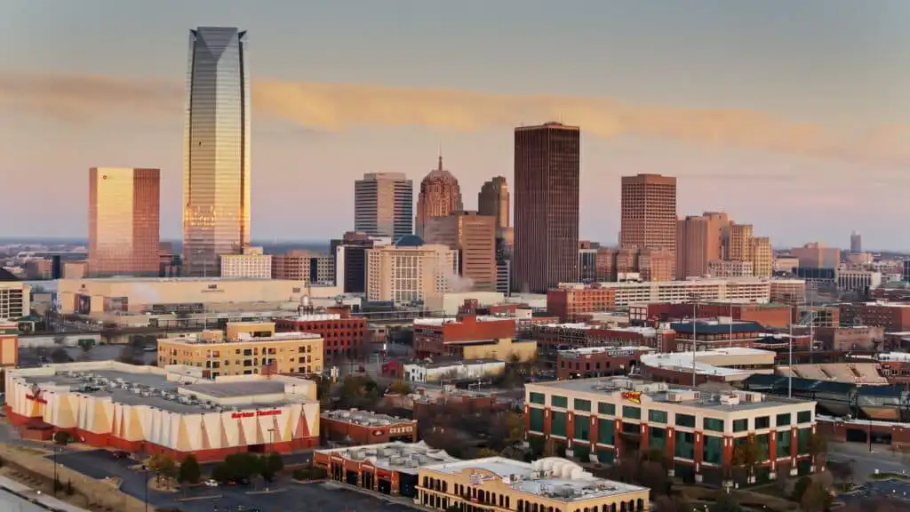 Downtown cityscape at sunset featuring tall skyscrapers, modern glass buildings, and a variety of smaller structures, with a clear sky and soft, warm light illuminating the scene.