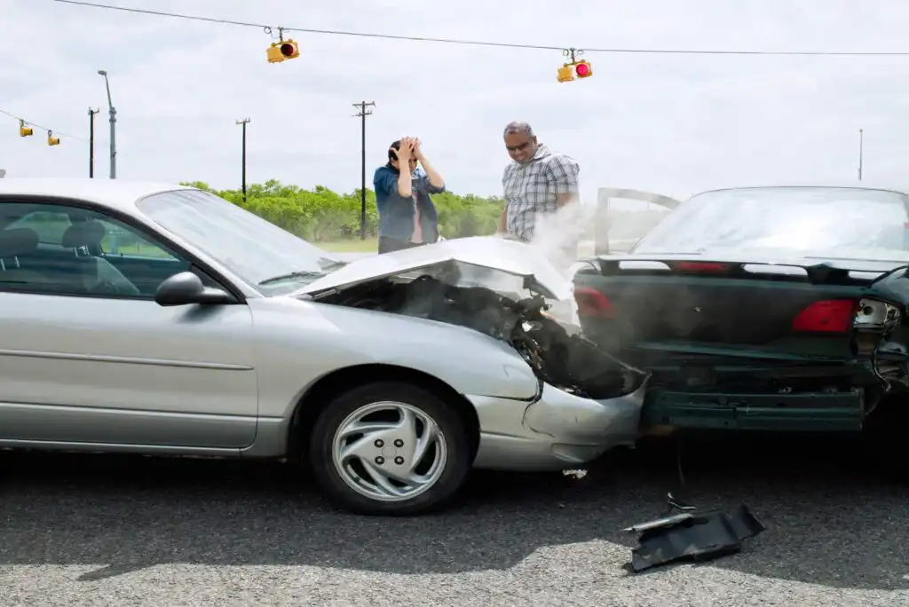 A silver car has collided with the rear of a dark green car at an intersection. Two people stand nearby, appearing distressed. Steam or smoke is coming from the damaged vehicles. Traffic lights hang overhead.