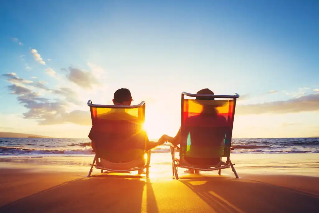 Two people sit in beach chairs on the sand, holding hands and facing a bright sunset over the ocean, with gentle waves and a partly cloudy sky.