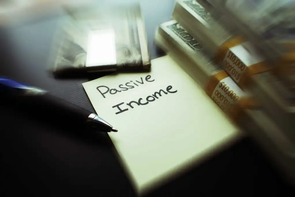 A pen and stacks of cash rest next to a yellow sticky note with Passive Income written on it, suggesting financial success through passive earnings.