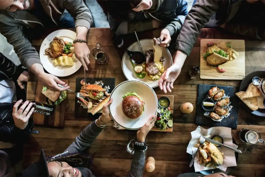 A group of people sitting around a wooden table, enjoying a variety of dishes such as burgers, salads, sushi, and omelets, seen from above. Hands reach for food and drinks, creating a lively, social atmosphere.
