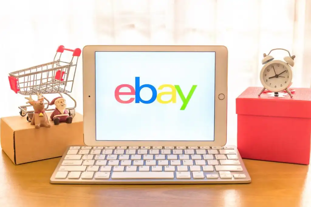 A tablet displaying the eBay logo is placed on a desk with a keyboard, a small shopping cart, a Santa figurine, a wrapped red gift box, and an alarm clock, all in front of a light curtain.