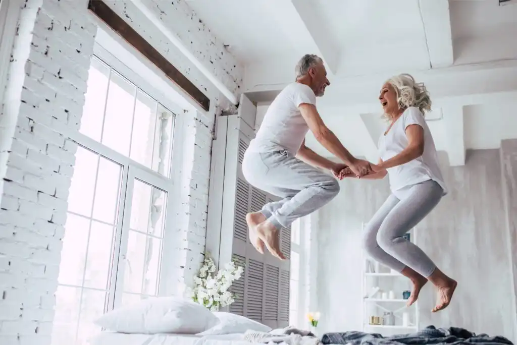 An older couple in matching white and gray clothes joyfully jump on a bed, holding hands and laughing in a bright, airy bedroom with large windows and white decor.