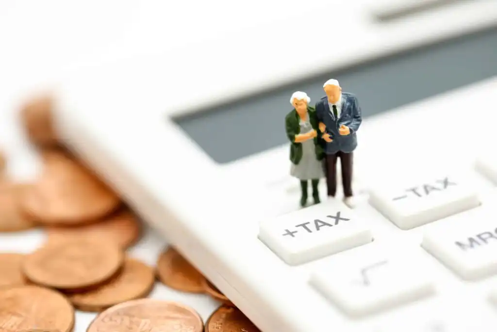 A miniature elderly couple stands on the tax buttons of a white calculator, surrounded by scattered pennies, symbolizing financial planning or tax issues for seniors.