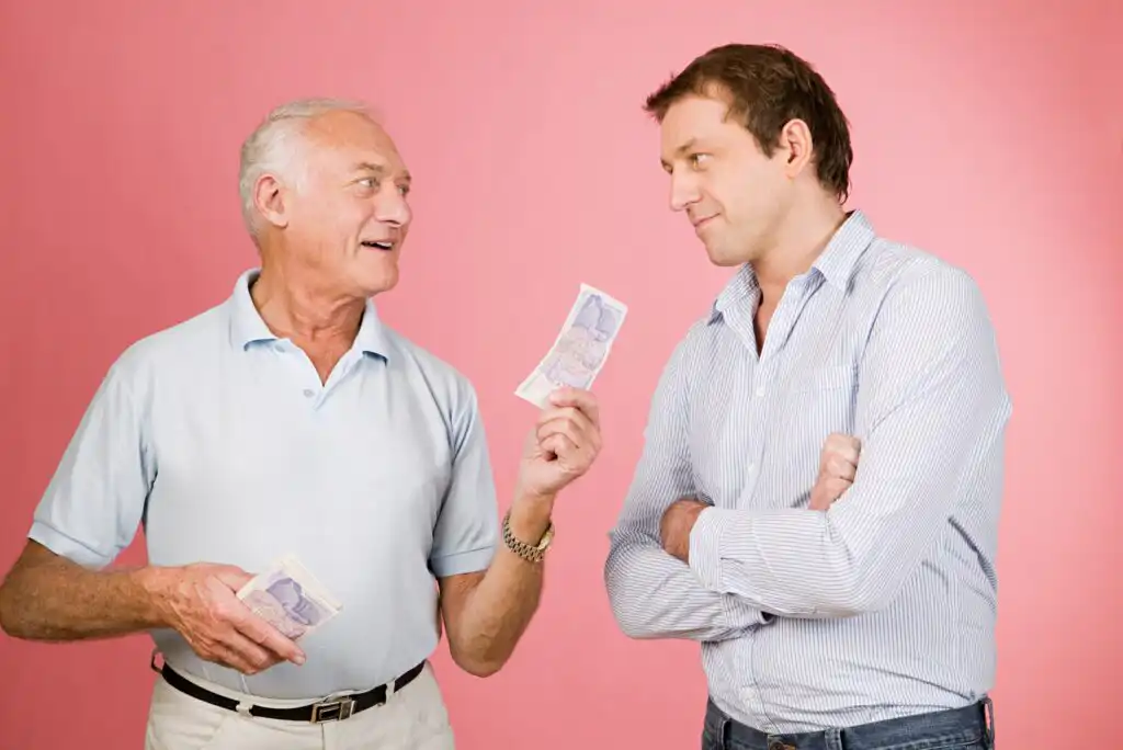 An older man smiling and handing cash to a younger man, who stands with his arms crossed and looks at him thoughtfully, both against a pink background.