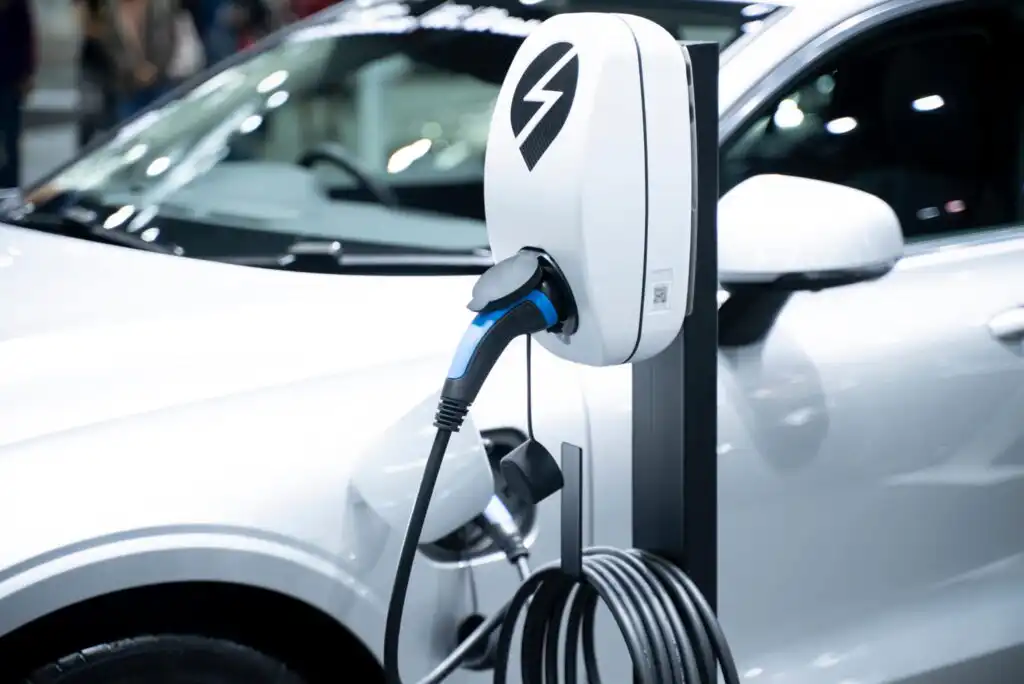 A white electric car is being charged at a charging station. The charging cable is plugged into the station, which displays a lightning bolt symbol, with the car’s side and wheel partially visible in the background.