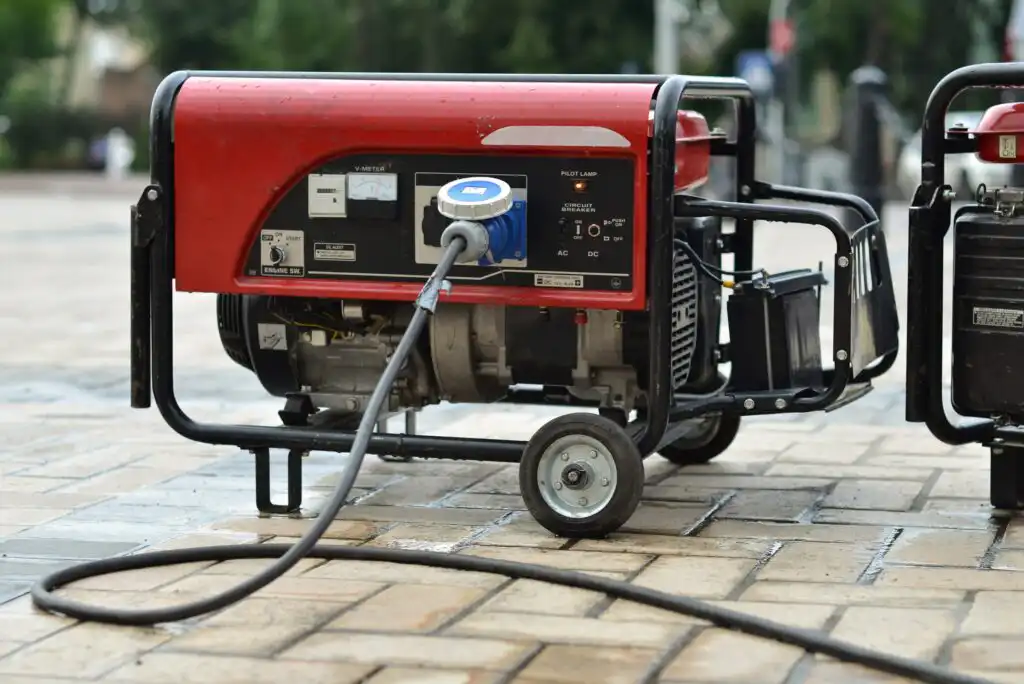A red portable generator with a control panel and attached cable sits on a paved outdoor surface. The generator has two wheels for mobility and is being used in an open, possibly public, area.
