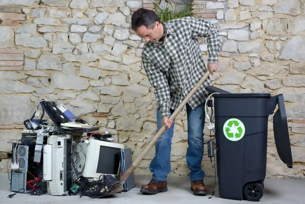 A man in a plaid shirt sweeps a pile of old electronics, including computers and cables, toward a black recycling bin with a green recycle symbol, against a stone wall background.