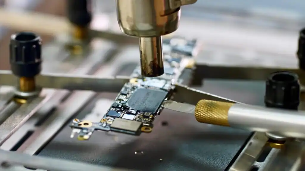 Close-up of a circuit board undergoing repair, with a heat gun above it and a precision tool placed nearby, highlighting the process of electronic component soldering or desoldering.