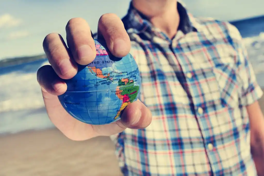 A person in a plaid shirt holds a small globe in their hand at the beach, with the ocean and sky in the background.