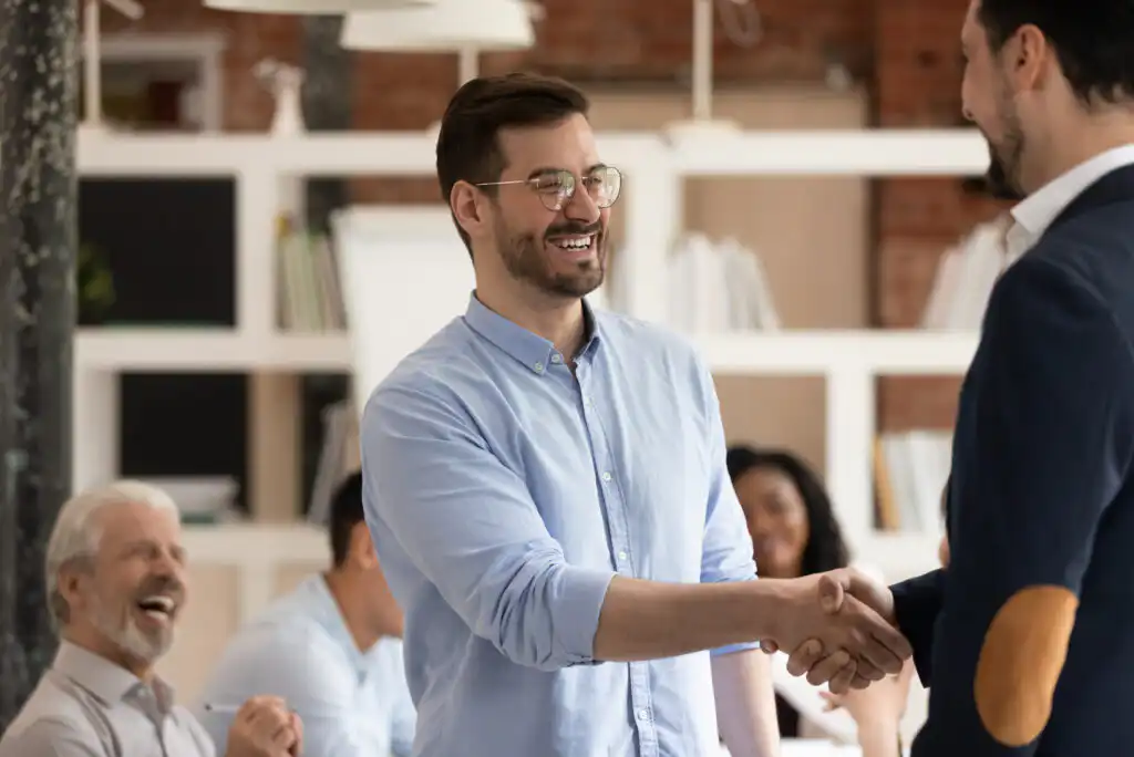 A smiling man in glasses and a light blue shirt shakes hands with another man in an office setting, while colleagues in the background appear happy and engaged.