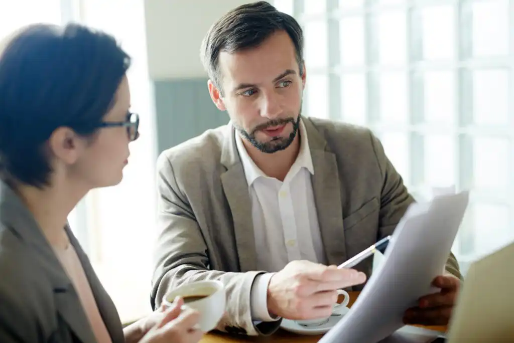A man in a suit explains a document to a woman with glasses who holds a cup of coffee. They are sitting together at a table, discussing paperwork in a bright, modern office setting.