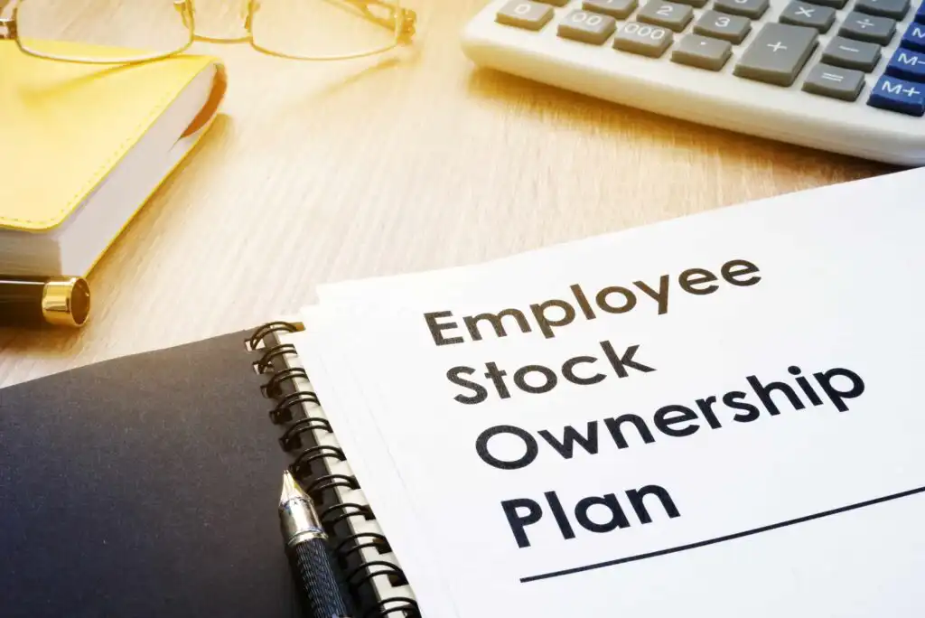 A spiral-bound notebook labeled Employee Stock Ownership Plan sits on a desk beside a calculator, a pair of eyeglasses, and a yellow-covered book.