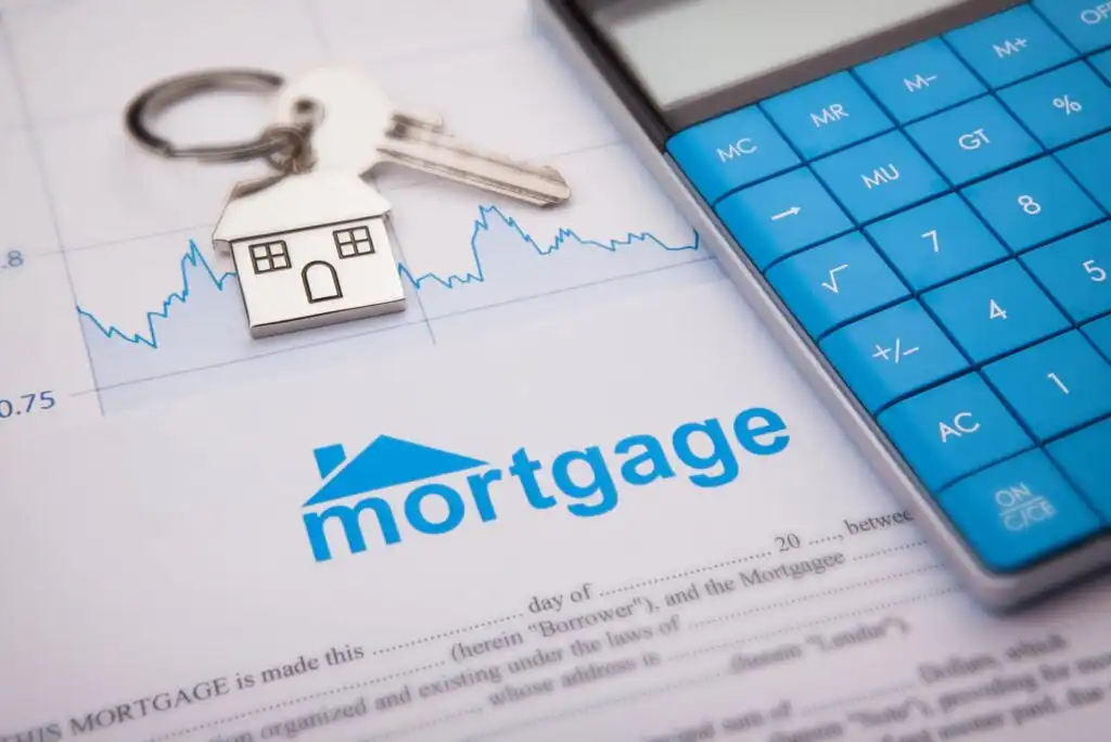 A set of house keys with a house-shaped keychain, a blue calculator, and a mortgage document on a desk, with a financial chart in the background.