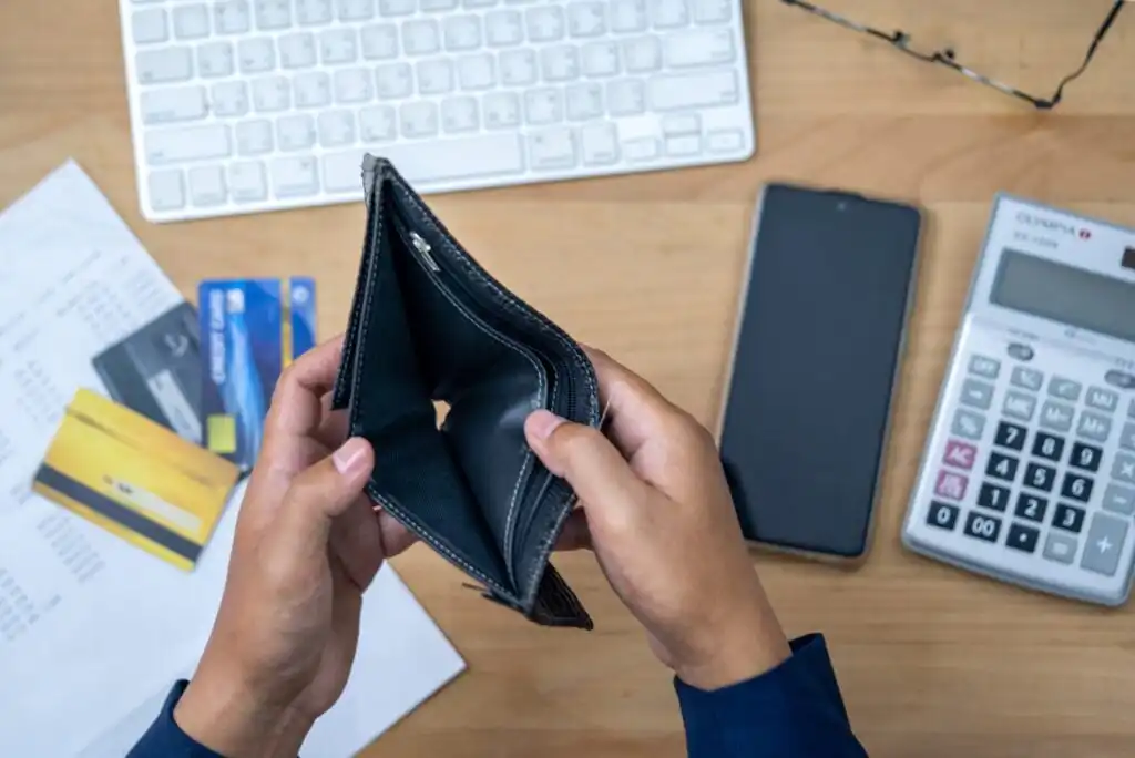 A person holds an open, empty wallet above a desk scattered with credit cards, papers, a keyboard, a calculator, and a smartphone.