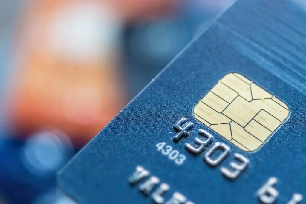Close-up image of a blue credit card with a visible gold chip and partial card number, showing fine details and texture. The background is blurred, highlighting the card’s surface and electronic chip.