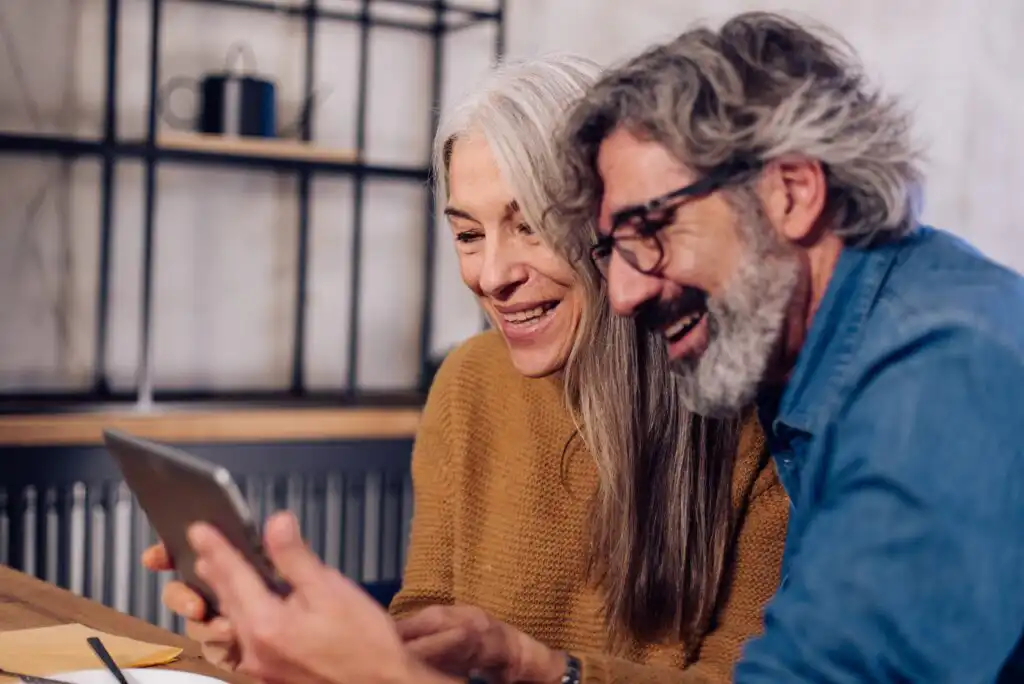An older couple with gray hair smiles and laughs while looking at a tablet together at a table indoors. The woman wears a mustard sweater; the man wears glasses and a blue shirt.