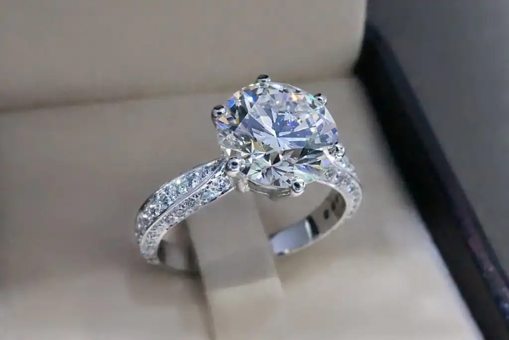 A close-up of a sparkling diamond engagement ring with a large round center stone and small diamonds set along the band, displayed in an open jewelry box.