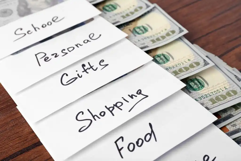 Several white envelopes labeled School, Personal, Gifts, Shopping, and Food are arranged in a row, each containing visible U.S. hundred-dollar bills sticking out on a wooden surface.