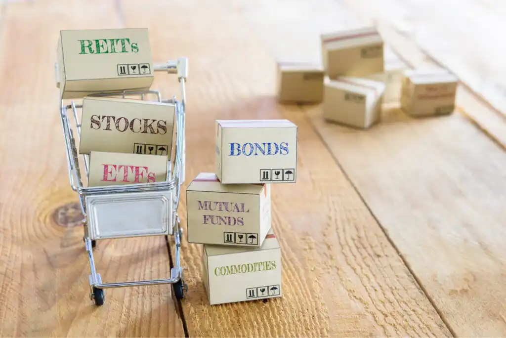 A miniature shopping cart filled with small boxes labeled REITs, Stocks, and ETFs, next to stacked boxes labeled Bonds, Mutual Funds, and Commodities on a wooden surface, symbolizing investment options.