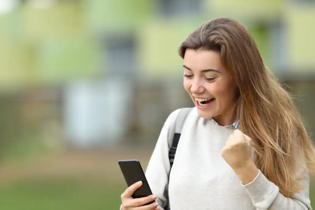 A young woman outdoors smiles excitedly at her phone, raising a fist in celebration. She has long hair, wears a light sweater, and carries a backpack. The background is blurred with green and yellow tones.