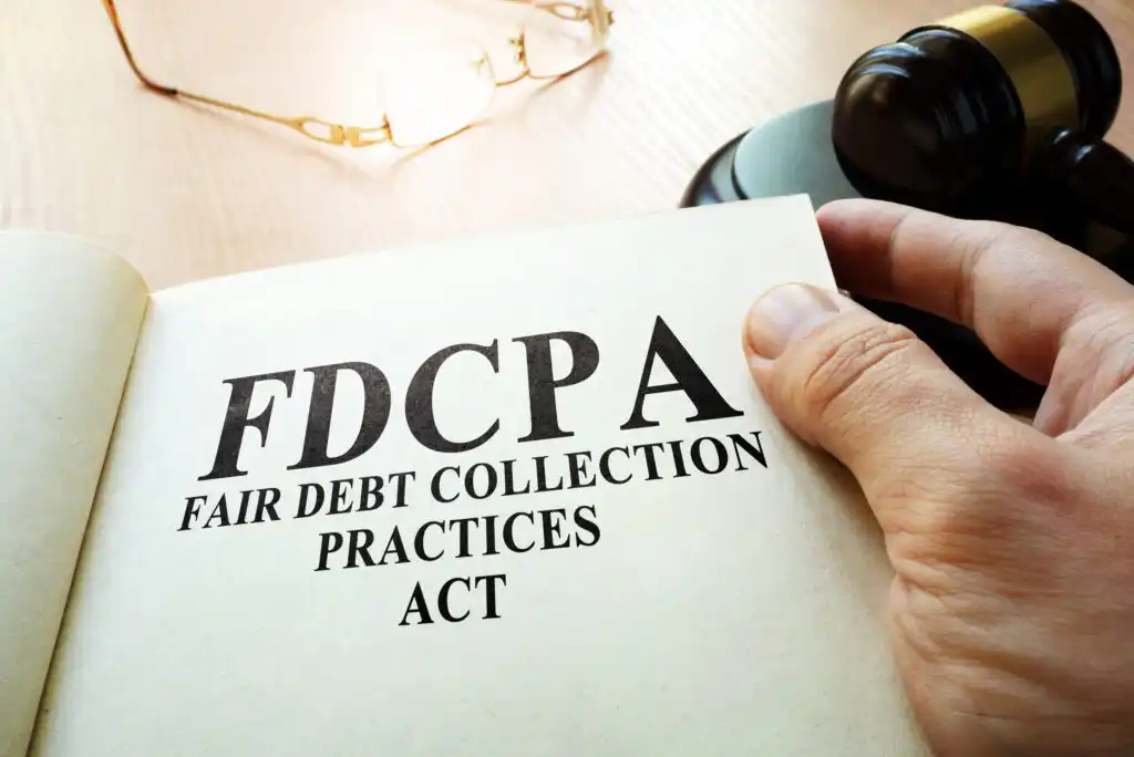 A person holds open a book displaying the text “FDCPA Fair Debt Collection Practices Act.” A judge’s gavel and eyeglasses are on the wooden desk in the background.