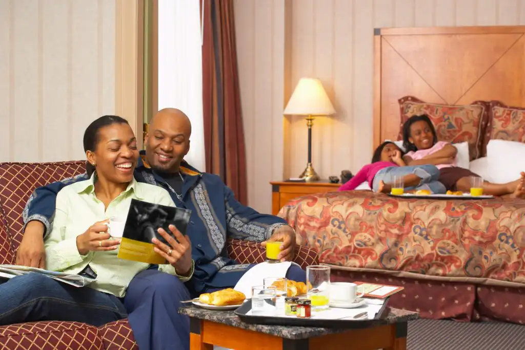 A smiling couple sits on a sofa drinking coffee and reading, while a woman and two children relax on a bed in the background, all in a cozy hotel room with breakfast on the table.