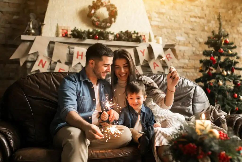 A smiling family of three sits on a couch holding sparklers, celebrating in a cozy living room decorated with a Christmas tree, wreath, and “Happy New Year” banner on the fireplace.