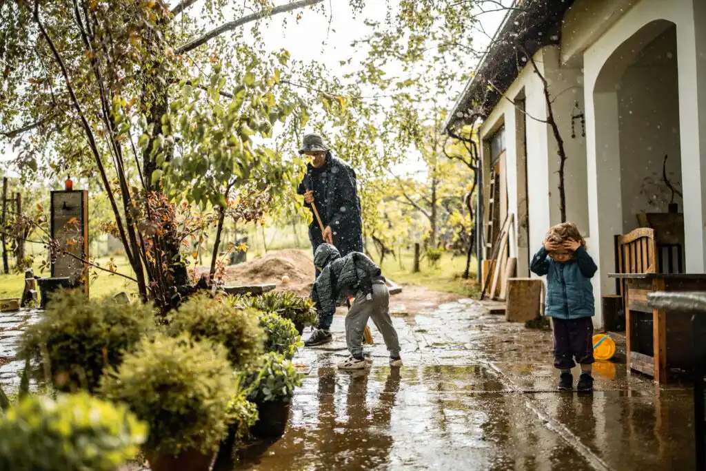 An adult and two children are outside near a house on a rainy day. The adult uses a shovel, one child bends down, and the other covers their face. Wet ground and lush plants are visible in the scene.