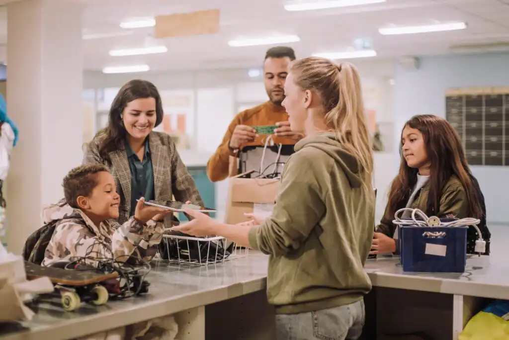 A group of people, including two adults and three children, gather at a counter, exchanging electronic items and cables in a bright, modern indoor space, suggesting a donation or recycling event.