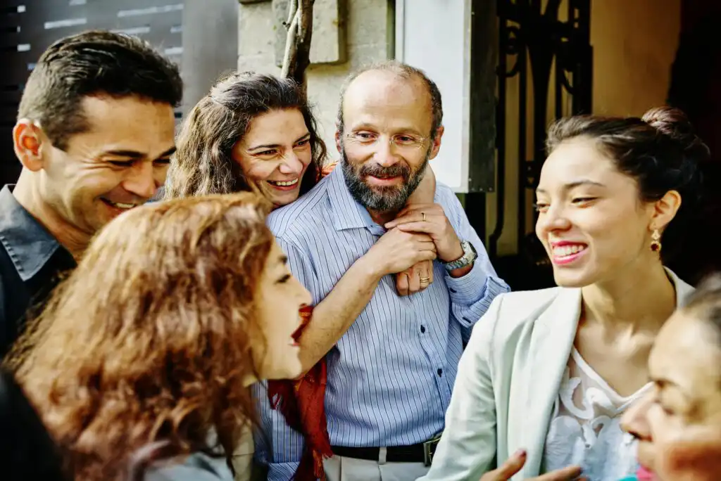 A group of people, including men and women, are gathered together, smiling and laughing outdoors. One woman stands behind a man, resting her hands on his shoulders, as others interact closely around them.
