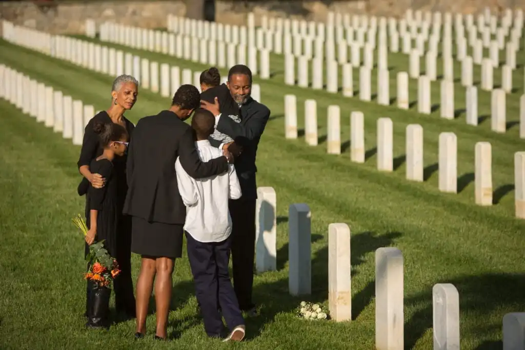 A family stands together, mourning at a cemetery with rows of white headstones. One man comforts a child, while a woman holds flowers. The group appears solemn, surrounded by green grass and gravestones.
