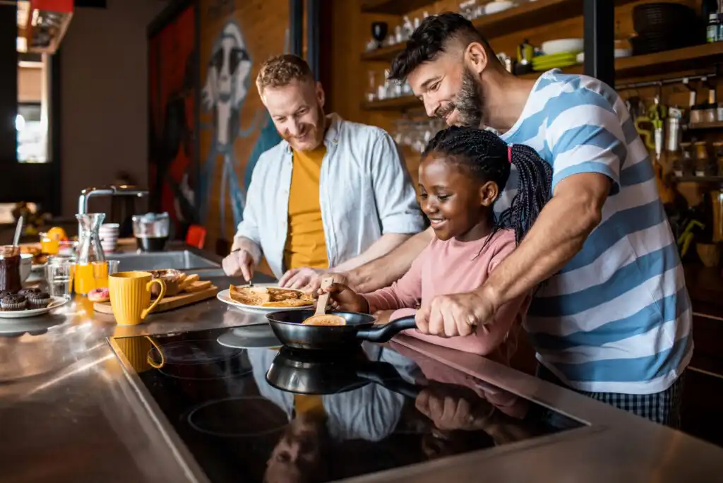 Two men and a young girl smile as they cook breakfast together in a modern kitchen. One man helps the girl flip food in a pan while the other prepares toast on a plate. Various kitchen items and food are visible on the counter.