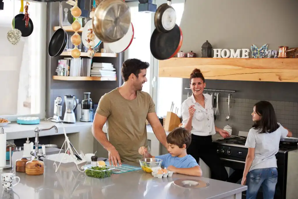 A smiling family of four prepares food together in a modern kitchen. The father and young son mix eggs at the counter, while the mother and daughter talk and cook near the stove. Pots and pans hang overhead.