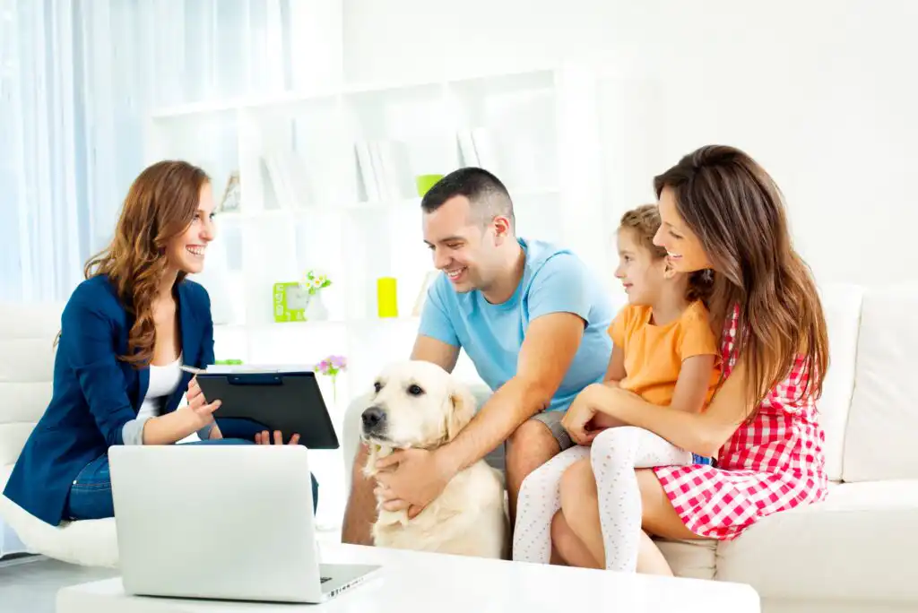 A woman with a clipboard talks to a smiling family—man, woman, girl, and their yellow labrador—sitting on a white couch in a bright living room. A laptop is on the table in front of them.
