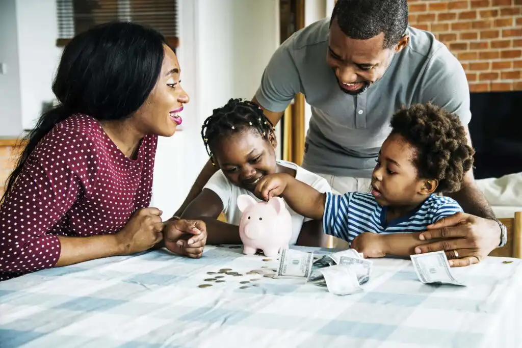 A smiling family of four sits at a table, watching two young children put coins in a pink piggy bank, with paper bills and coins scattered on the table.